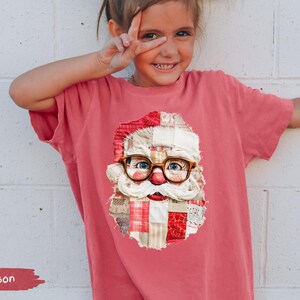 May include: Crimson-coloured t-shirt featuring a Santa Claus graphic with glasses and a patchwork design. The shirt is worn by a young person with white bows in their hair. The word "Crimson" is displayed on the bottom left.