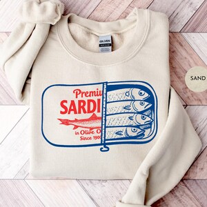 May include: Sand-colored sweatshirt featuring a graphic of a sardine can with the words "Premium Sardi" in red and blue. The design includes illustrations of sardines and the text "in Olive Oil Since 1900".