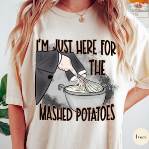 Puede incluir: Camiseta color crema con el texto "I'M JUST HERE FOR THE MASHED POTATOES". El gráfico muestra una mano con traje removiendo puré de papas con un batidor. La camiseta es de corte holgado.