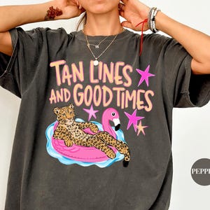 May include: A charcoal grey short-sleeve t-shirt featuring a graphic print of a leopard lounging on a pink flamingo pool float. The text reads "TAN LINES AND GOOD TIMES" in peach-coloured letters, accented by several pink and yellow stars.