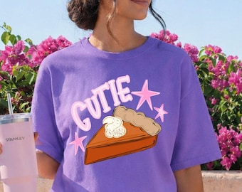 May include: A violet t-shirt featuring a graphic of a slice of pumpkin pie with whipped cream, the text "CUTIE", and pink stars. The shirt is paired with a matching violet baseball cap and a light violet insulated tumbler with a straw.