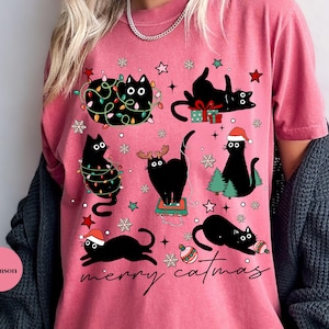 May include: Crimson-colored t-shirt featuring a playful design of black cats decorated with Christmas lights, Santa hats, and ornaments. The phrase "merry catmas" is written in a cursive font. The shirt has a relaxed fit.