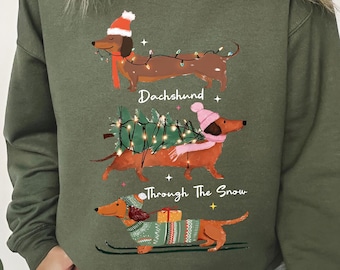 Peut inclure: Sweat-shirt vert militaire avec un motif de teckels festifs. Trois teckels sont représentés : un avec un bonnet de Père Noël, un portant un sapin de Noël et un faisant du ski avec un cadeau. Le texte dit "Dachshund Through The Snow."