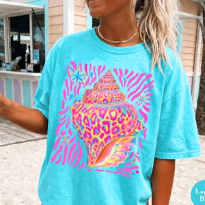 May include: A turquoise t-shirt featuring a seashell design with pink, yellow, and blue accents. The seashell has a leopard print pattern. The shirt is labeled "Lagoon Blue".