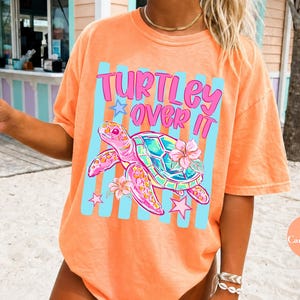 May include: Orange t-shirt with the text "Turtley Over It" and a graphic of a sea turtle with flowers. The shirt has a relaxed fit and short sleeves. The design includes blue stripes and pink stars. The shirt is a vibrant, summery style.
