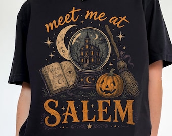 May include: A black short-sleeve t-shirt featuring a graphic print with the text "meet me at SALEM". The design includes a crystal ball with a house inside, an open spellbook, a pumpkin, a broomstick, and a crescent moon in orange and brown tones.