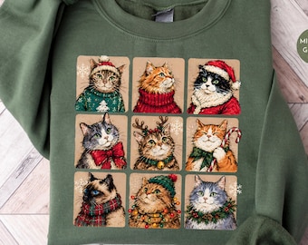May include: A military green crewneck sweatshirt featuring a grid of nine illustrations of cats dressed in holiday attire, including Santa hats, scarves, sweaters, and festive decorations like candy canes and lights, with a small circular label.