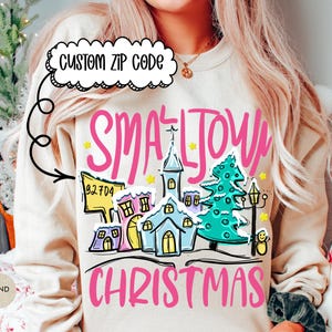 May include: Sand-colored sweatshirt with a colorful graphic of a small town Christmas scene. The design includes houses, a church, a Christmas tree, and the words "Small Town Christmas" in pink. A custom zip code is also featured.