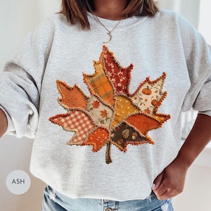 Peut inclure: Un sweat-shirt à col rond gris cendré arborant un grand motif central en forme de feuille d'érable. La feuille est composée de divers motifs de tissu style patchwork dans des tons orange, marron et crème, incluant vichy et fleurs.