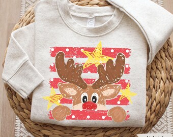 May include: A natural-colored crewneck sweatshirt featuring a graphic print of a reindeer with brown antlers and a red nose. The design includes red horizontal stripes with white polka dots and yellow stars, rendered in a textured, crayon-like style.