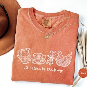 May include: A coral-colored t-shirt with a white graphic of a pitcher, teacup, spoons, and a hen in a basket. The text "I'd rather be thrifting" is below the graphic. A gold necklace is draped on the shirt.