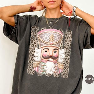 Può includere: Una t-shirt a maniche corte grigio antracite con una grafica centrale di un soldatino schiaccianoci che fa l'occhiolino. Lo schiaccianoci indossa un'uniforme con stampa leopardata rosa e bianca con dettagli dorati su sfondo leopardato rettangolare.