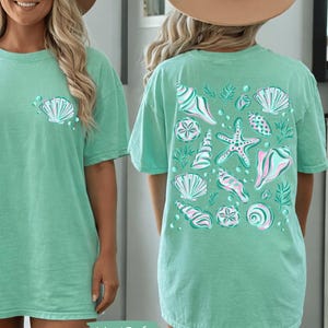 May include: A mint green t-shirt with a seashell graphic on the front and back. The back of the shirt features a collection of seashells, starfish, and aquatic plants in shades of white, pink, and teal. The text "Island Reef" is at the bottom.