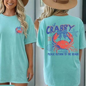 Puede incluir: Una camiseta turquesa con un gr&aacute;fico de cangrejo. La parte delantera presenta un peque&ntilde;o dise&ntilde;o de cangrejo, mientras que la parte trasera muestra una ilustraci&oacute;n de cangrejo m&aacute;s grande con el texto "If Crabby, Please Return to the Beach". La camiseta es de corte holgado.