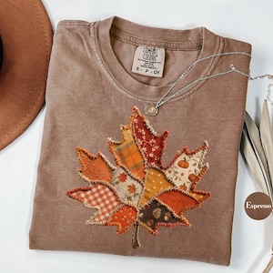 Könnte beinhalten: Ein braunes Kurzarm-T-Shirt mit einem Comfort Colors-Etikett und einer großen Ahornblatt-Applikation auf der Vorderseite. Das Blatt besteht aus verschiedenen Stoffstücken mit orangefarbenen, roten und gelben Mustern.