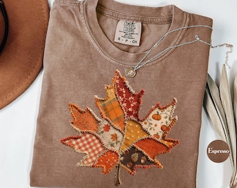 May include: A brown short-sleeve t-shirt with a Comfort Colors label and a large maple leaf applique on the front. The leaf is constructed from various fabric patches featuring orange, red, and yellow patterns, including pumpkins and gingham.