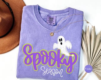 May include: A violet short-sleeved t-shirt with a crew neckline featuring a white ghost graphic and the purple text Spooky Season. The shirt has a Comfort Colors label at the inner neck and is displayed with a brown hat and a gold chain necklace.