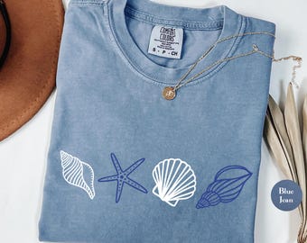 Op de afbeelding: Een jeansblauw t-shirt met een wit schelpje, een blauwe zeester en een blauwe schelp. De tekst "Comfort Colors" is zichtbaar op het label.