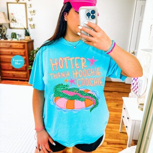 Puede incluir: Camiseta azul laguna con el texto "Hotter Than a Hoochie Coochie" en naranja y rosa. La camiseta presenta un gr&aacute;fico de un caim&aacute;n verde flotando en una boya rosa y naranja.