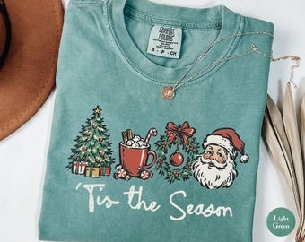 May include: A light green short-sleeve t-shirt featuring a graphic print of a Christmas tree, a mug of hot cocoa, a wreath, and Santa Claus. The text 'Tis the Season' is printed in white below the graphics. The shirt has a Comfort Colors label.
