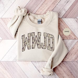 May include: A sand-colored sweatshirt with the letters "WWJD" in a camouflage pattern across the chest. The sweatshirt has a crew neck and long sleeves. The word "SAND" is printed on a small, round sticker.