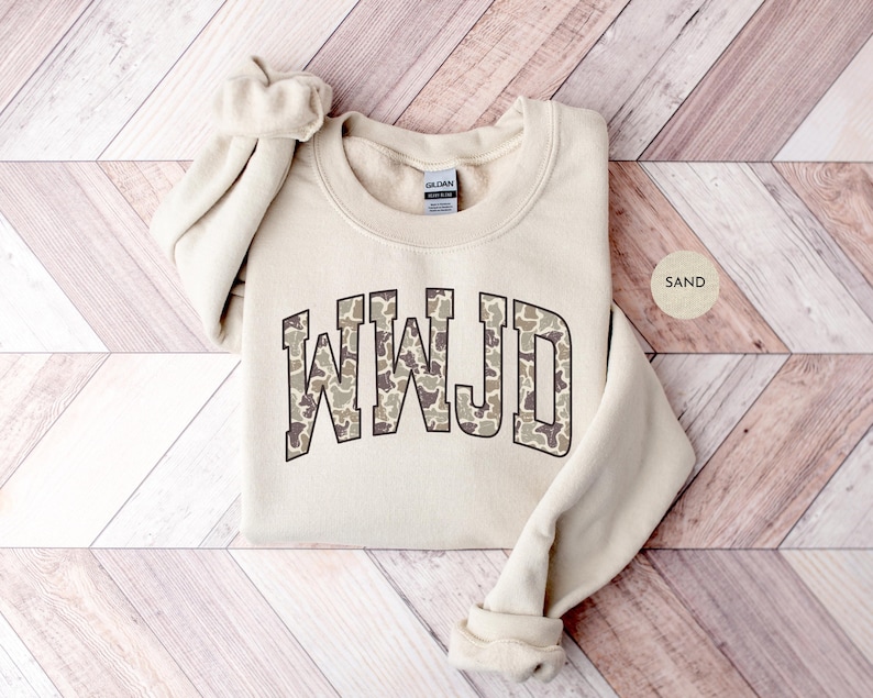 May include: A sand-colored sweatshirt with the letters "WWJD" in a camouflage pattern across the chest. The sweatshirt has a crew neck and long sleeves. The word "SAND" is printed on a small, round sticker.