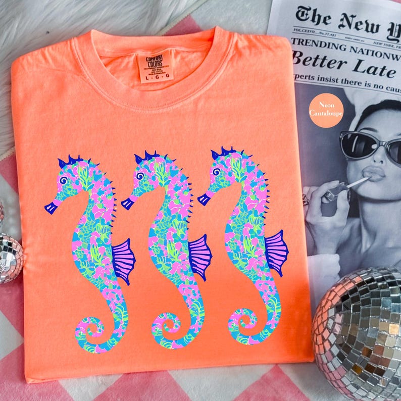 May include: Neon orange t-shirt featuring three seahorse designs. The seahorses are decorated with pink, blue, and green floral patterns. The shirt has a crew neck and short sleeves. The tag reads "Comfort Colors".