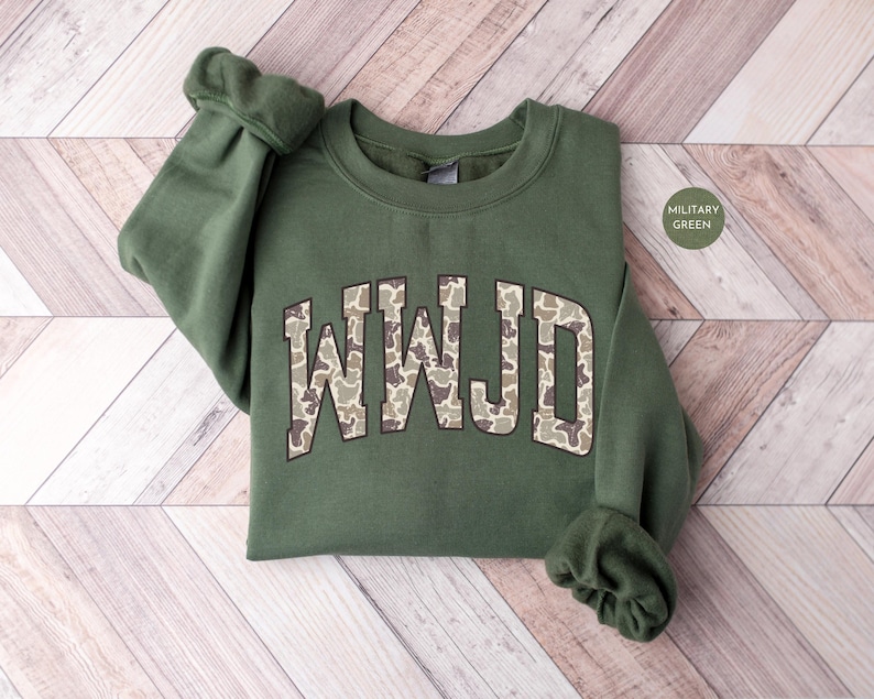 May include: A military green sweatshirt with the letters "WMJD" in a camouflage pattern across the chest. The sweatshirt is made of a soft material and has a crew neck and long sleeves. The words "MILITARY GREEN" are in a circle in the upper right corner.