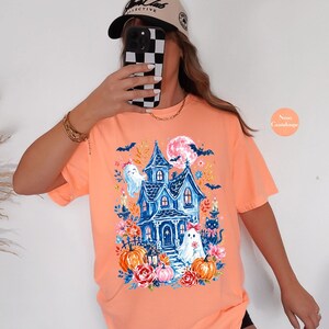 K&ouml;nnte beinhalten: Ein pfirsichfarbenes T-Shirt mit einem Grafikdruck eines blauen Spukhauses, zwei wei&szlig;en Geistern mit rosa Schleifen, einer schwarzen Katze, orangefarbenen K&uuml;rbissen, rosa Blumen, Flederm&auml;usen und einem Vollmond.