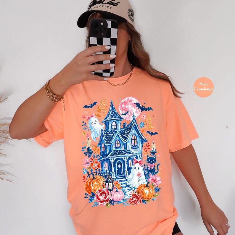 K&ouml;nnte beinhalten: Ein pfirsichfarbenes T-Shirt mit einem Grafikdruck eines blauen Spukhauses, zwei wei&szlig;en Geistern mit rosa Schleifen, einer schwarzen Katze, orangefarbenen K&uuml;rbissen, rosa Blumen, Flederm&auml;usen und einem Vollmond.