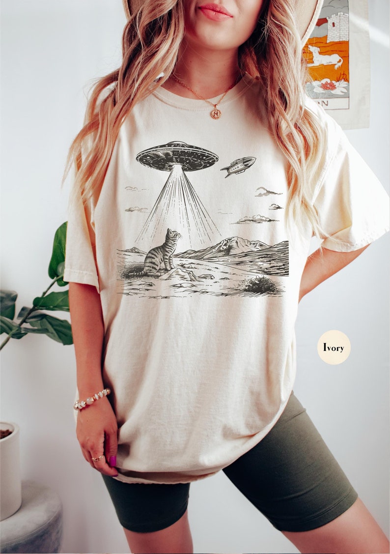 Może przedstawiać: Kobieta w kremowej koszulce z czarno-białym nadrukiem kota patrzącego w g&oacute;rę na UFO. UFO emituje wiązki światła. Tekst "Kremowy" jest widoczny w prawym dolnym rogu obrazu.
