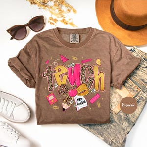 May include: Espresso-colored t-shirt with the word "TEACH" in a playful design. The letters have a leopard print and pink accents, with school-themed illustrations like an apple, pencil, and paper airplane. Sunglasses, a hat, and jeans are also in the image.