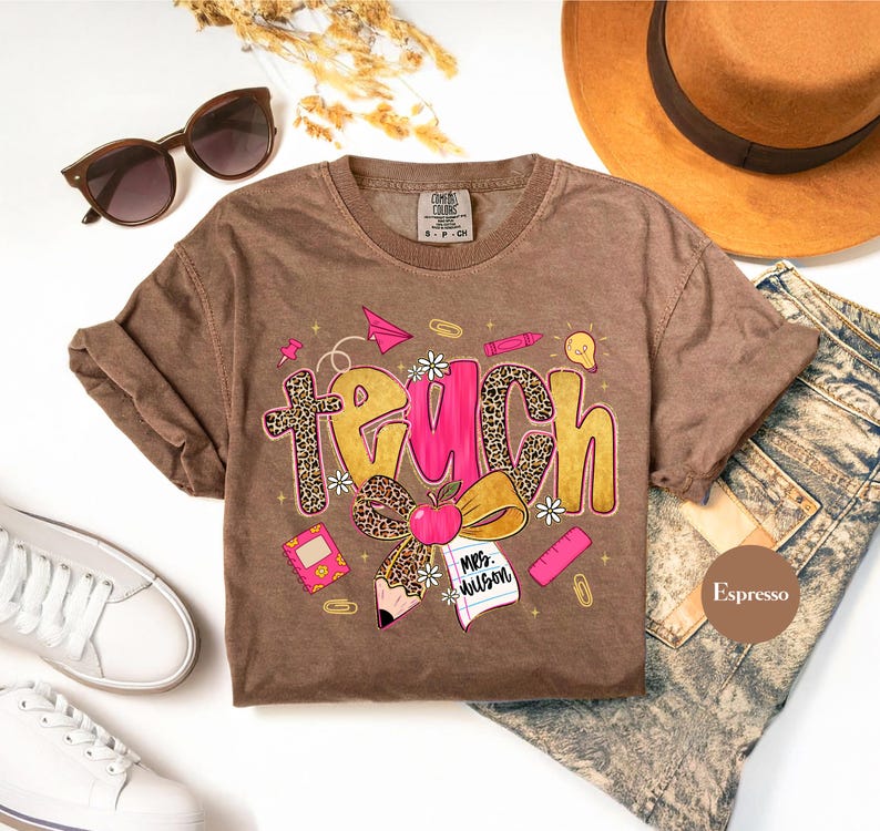 May include: Espresso-colored t-shirt with the word "TEACH" in a playful design. The letters have a leopard print and pink accents, with school-themed illustrations like an apple, pencil, and paper airplane. Sunglasses, a hat, and jeans are also in the image.