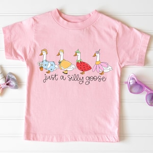 以下が含まれることがあります： ピンクの半袖Tシャツに、異なるコスチュームを着た5羽のガチョウのグラフィックがプリントされています。ガチョウの下には「just a silly goose」というテキストがプリントされています。