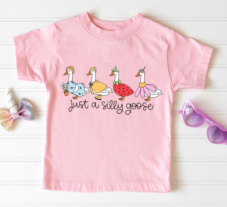 以下が含まれることがあります： ピンクの半袖Tシャツに、異なるコスチュームを着た5羽のガチョウのグラフィックがプリントされています。ガチョウの下には「just a silly goose」というテキストがプリントされています。
