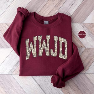 May include: Maroon-colored sweatshirt with the letters "WWJD" in a camouflage pattern across the chest. The sweatshirt is folded on a light-colored wooden surface. A maroon-colored circle with the word "MAROON" is in the upper right corner.