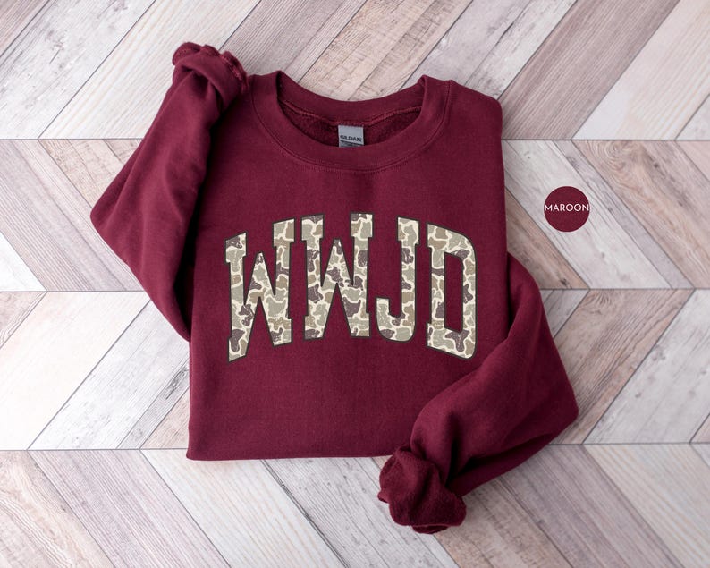May include: Maroon-colored sweatshirt with the letters "WWJD" in a camouflage pattern across the chest. The sweatshirt is folded on a light-colored wooden surface. A maroon-colored circle with the word "MAROON" is in the upper right corner.