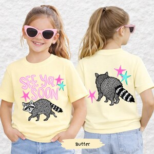 May include: A light yellow t-shirt featuring a raccoon graphic and the text "See Ya Soon" in pink and teal. The back of the shirt also displays a raccoon and star graphics. The shirt is made of a soft material.