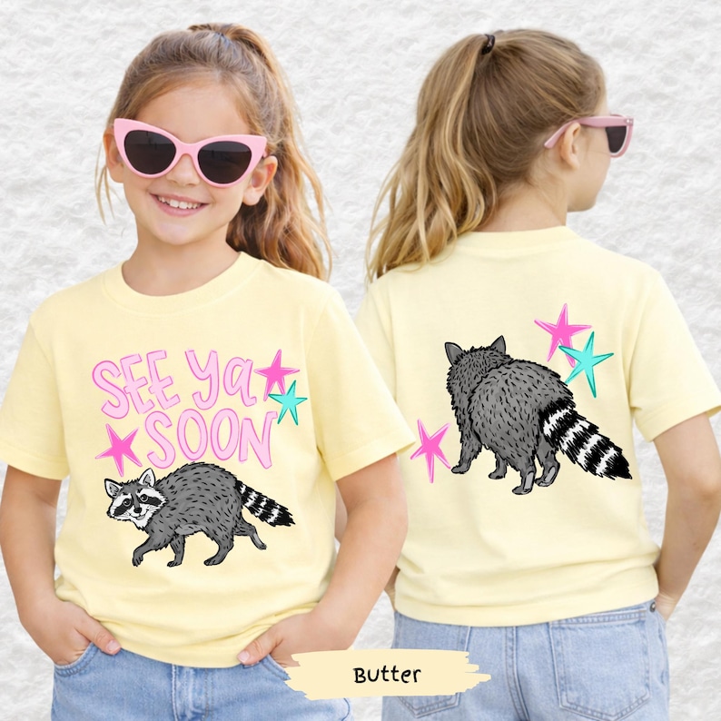 May include: A light yellow t-shirt featuring a raccoon graphic and the text "See Ya Soon" in pink and teal. The back of the shirt also displays a raccoon and star graphics. The shirt is made of a soft material.