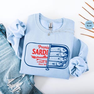 May include: Light blue sweatshirt with a graphic of a sardine can. The can design features the words "Premium Sardi" in red, a red fish illustration, and the text "in Olive Oil Since 1900". The sweatshirt is paired with blue jeans.