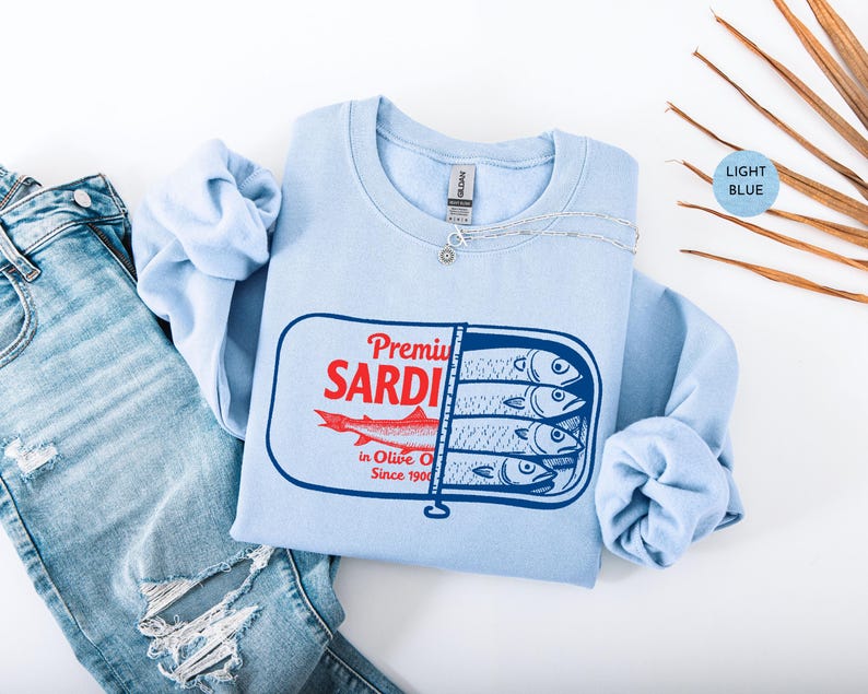 May include: Light blue sweatshirt with a graphic of a sardine can. The can design features the words "Premium Sardi" in red, a red fish illustration, and the text "in Olive Oil Since 1900". The sweatshirt is paired with blue jeans.