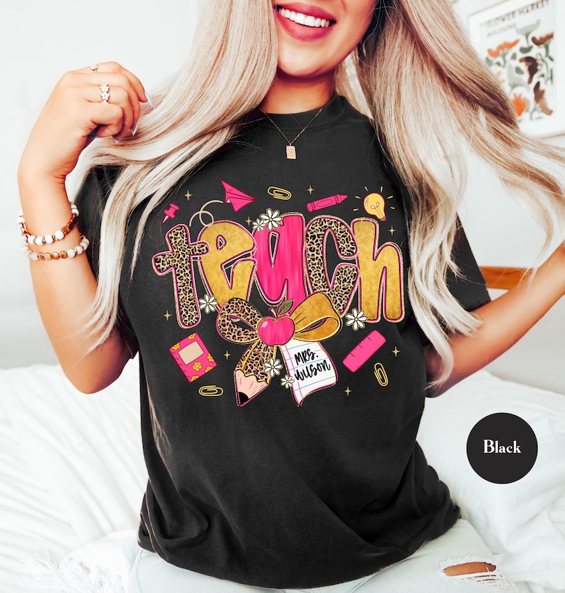 May include: Black t-shirt with the word "Teach" in a playful design. The word "Teach" features a leopard print pattern, pink and gold colors, and school-themed graphics like an apple, pencil, and paper airplane.