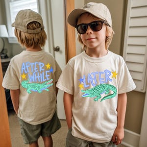 Puede incluir: Dos ni&ntilde;os con camisetas color crema con un dise&ntilde;o de cocodrilo de dibujos animados. Una camiseta dice "AFTER WHILE" y la otra "LATER" en letras azules con burbujas. Ambas camisetas tienen un cocodrilo verde azulado y estrellas amarillas y azules.