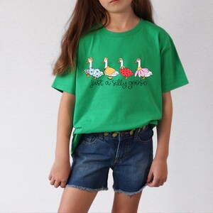 以下が含まれることがあります： さまざまな衣装を着たガチョウの列と「Just a silly goose」というテキストが書かれた緑色のTシャツ。モデルはデニムのショートパンツを履いています。