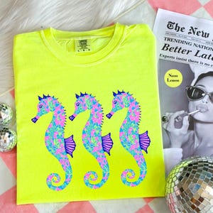 May include: Neon yellow t-shirt featuring three seahorse designs in pink, blue, and purple. The shirt has a crew neck and short sleeves. The text "Neon Lemon" is visible on a newspaper in the background.