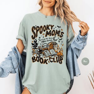 May include: A sage green t-shirt featuring a graphic print with the text "SPOOKY MOMS BOOK CLUB" and "Running on coffee, horror & chapters". The design includes illustrations of stacked books, a potted plant, candles, a pumpkin, and bats.