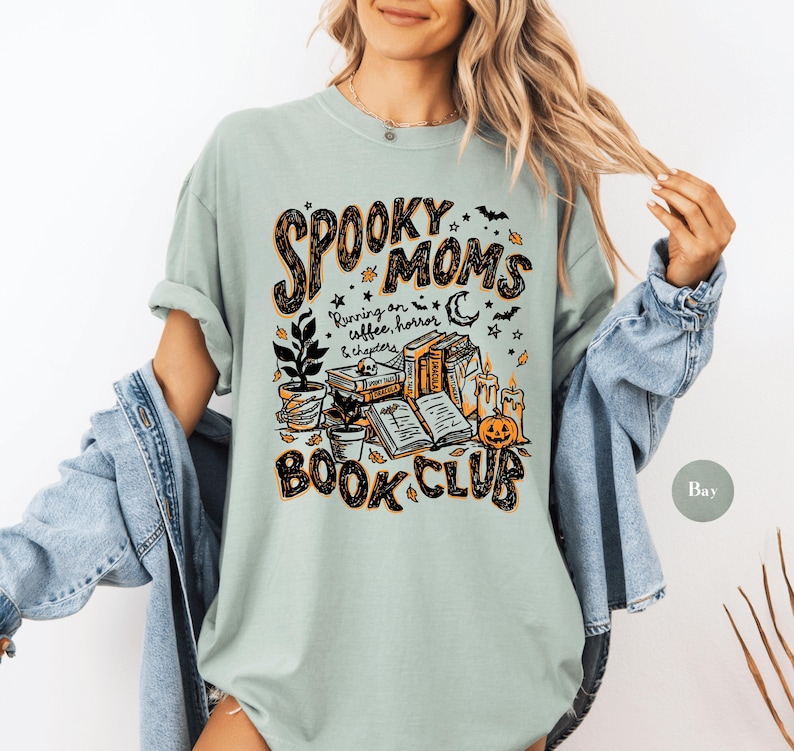 May include: A sage green t-shirt featuring a graphic print with the text "SPOOKY MOMS BOOK CLUB" and "Running on coffee, horror & chapters". The design includes illustrations of stacked books, a potted plant, candles, a pumpkin, and bats.