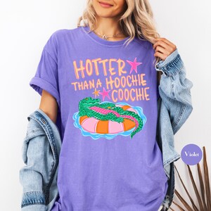 Puede incluir: Camiseta violeta con la frase "Hotter Than a Hoochie Coochie" en rosa y amarillo. El gr&aacute;fico presenta un caim&aacute;n verde sobre un flotador inflable naranja y rosa. La camiseta tiene mangas enrolladas.