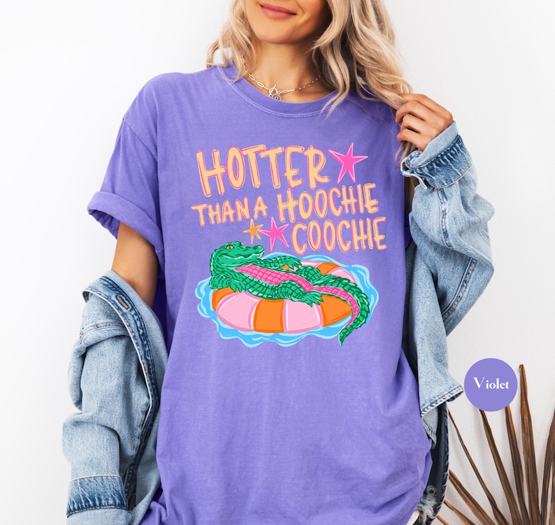 Puede incluir: Camiseta violeta con la frase "Hotter Than a Hoochie Coochie" en rosa y amarillo. El gr&aacute;fico presenta un caim&aacute;n verde sobre un flotador inflable naranja y rosa. La camiseta tiene mangas enrolladas.