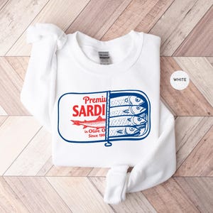 May include: White sweatshirt featuring a graphic of a sardine can with the words "Premium Sardines in Olive Oil Since 1900". The sardine can design is in red and blue. The sweatshirt is white.